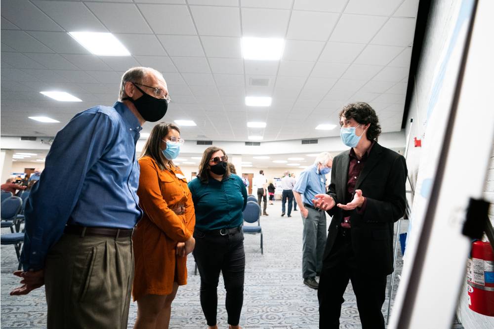 Scholar giving poster presentation to three guests, all wearing masks.